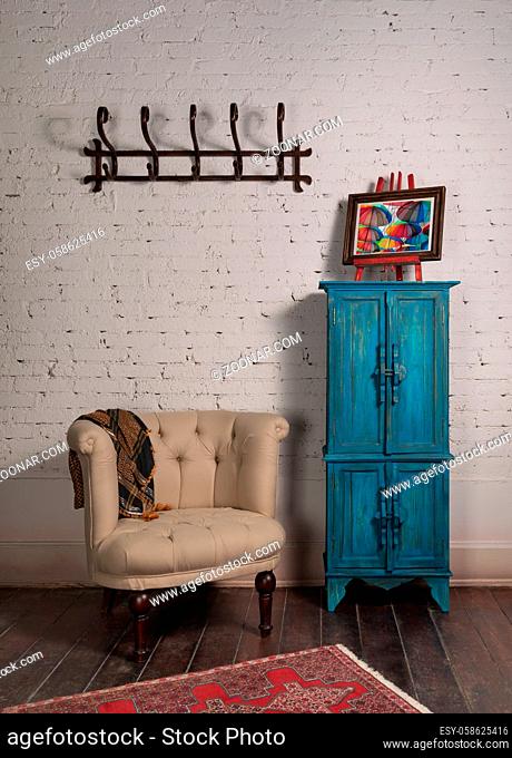Classic beige armchair with wooden legs, vintage turquoise cupboard, wall hanger, and ornate scarf on white bricks wall and grunge wooden parquet with red...