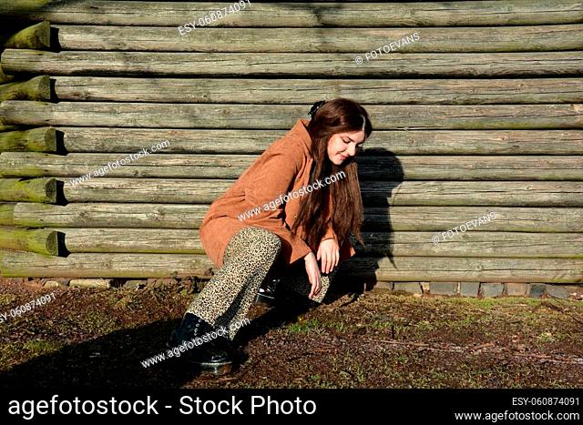 Young girl squats on the floor in front of old wooden planks, in the sunlight and looks down