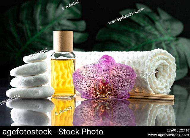 Beautiful lilac orchid flower, clear bottle of yellow oil or perfume, wooden sticks and rolled towel with stack of white stones and monstera leaves