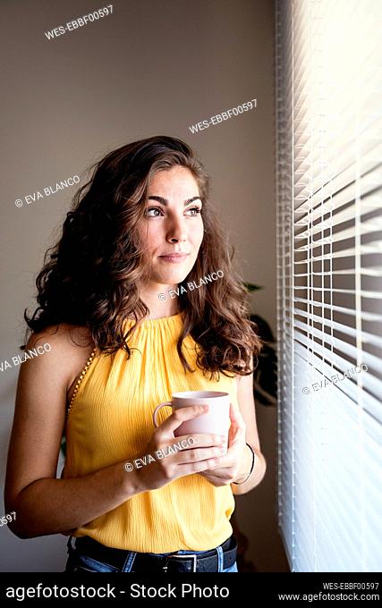 Thoughtful woman holding coffee cup while looking away through blinds at home
