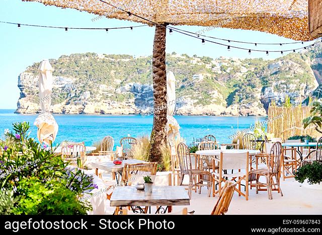 Interior of coastal beach restaurant. Under canopy located empty wooden tables and chairs, cozy cafe with beautiful view to rocky mountains hillside village and...