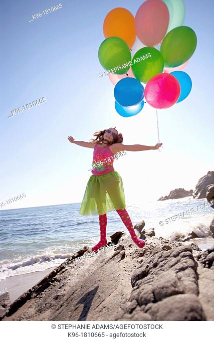 little girl holding balloons and jumping while on the rocks at the ocean