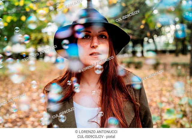 Young woman with long red hair amongst floating bubbles in autumn park, shallow focus portrait