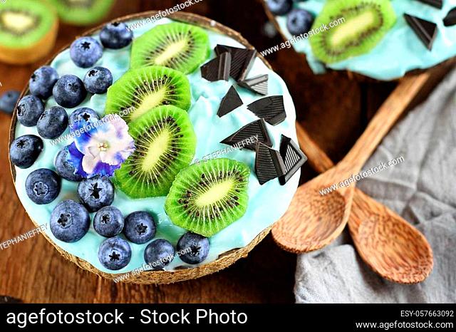 Blue spirulina and berry smoothie bowl, fresh blueberries, kiwi and chocolate pieces with wooden spoons served in coconut bowls over a rustic background