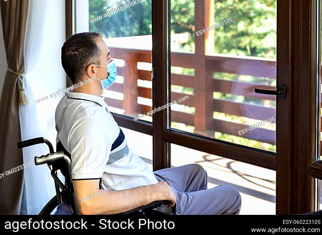 Lonely young man wearing face mask sitting in a wheelchair alone looking out the window. Focus on his face