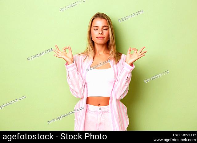 Portrait of young relaxed woman with blond short hairstyle, closing eyes and holding hands in zen gesture, meditating, practice yoga over green background