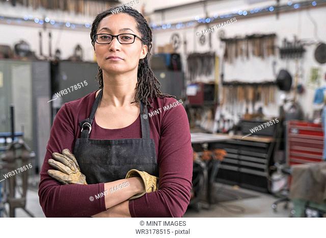 Woman with brown hair wearing glasses and apron standing in metal workshop, arms folded, looking at camera