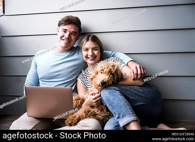 Happy young couple sitting with dog in front of wall