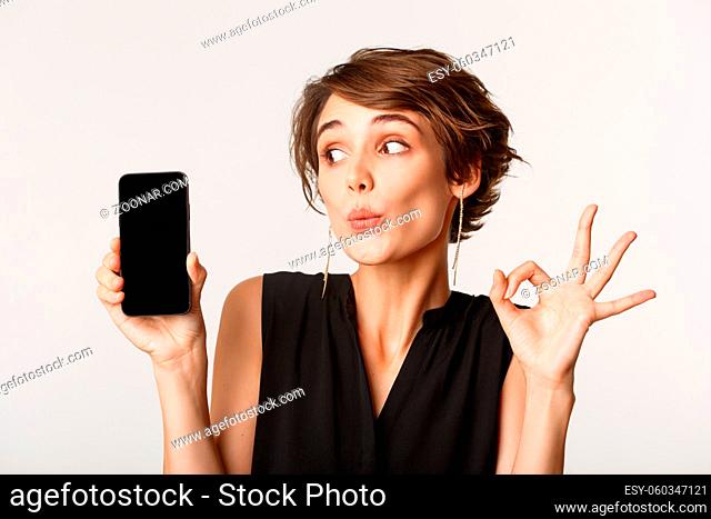 Close-up of silly pretty woman pouting, showing mobile phone screen and okay gesture, standing over white background