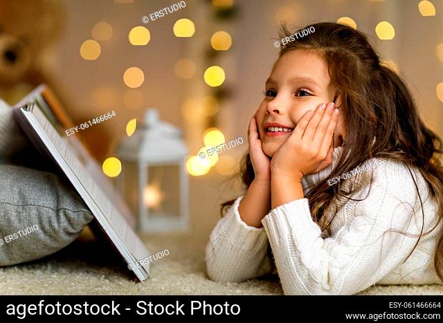 Happy smiling little girl is reading fairy tale book on the background with lights. child dreams of a wonderful christmas