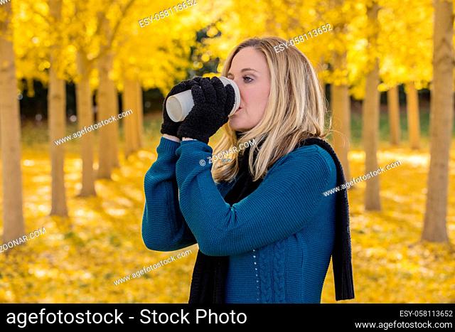 A beautiful blonde model drinking a hot beverage while posing outdoors in a field of yellow leaves