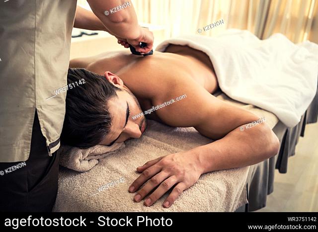 Young man relaxing during traditional massage with hot stones at Asian spa and wellness center