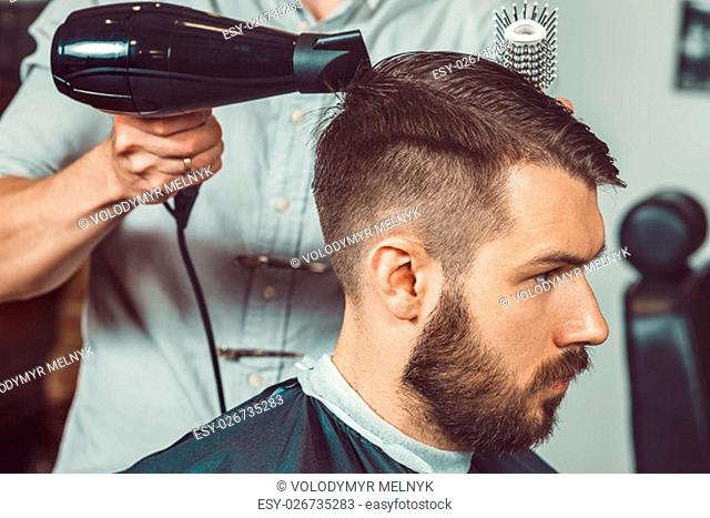 The hands of young barber making haircut of attractive bearded man in barbershop