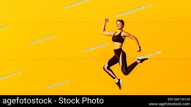 Side view of young satisfied attractive sporty woman in fashionable black sportswear runner in on orange wall background. Dynamic jumping movement