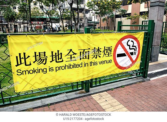 Hong Kong, China, Asia. Hong Kong Kowloon. Large yellow bilingual banner in english and chinese at the entrance to a small urban park advising that ""Smoking is...