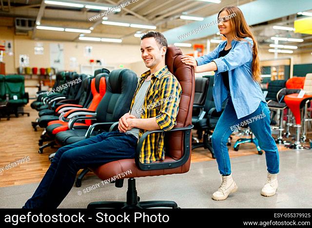 Happy young couple rides on office chair in furniture store showroom. Man and woman looking samples for bedroom in shop, husband and wife buys goods for modern...