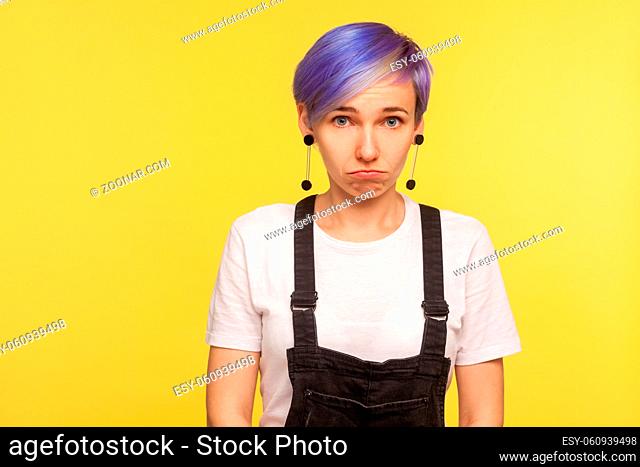 Portrait of young upset hipster woman with violet dyed short hair in denim overalls looking at camera with resentful offended expression, pursed lips