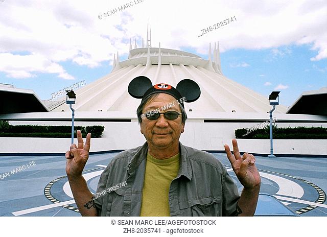 An asian man holds peaces signs while wearing a Mickey Mouse cap in front of Space Mountain in Disneyland