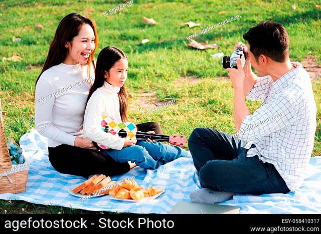 Happy Asian young family father, mother and child having fun and enjoying outdoor together sitting on the grass party with shooting photos by retro camera a...