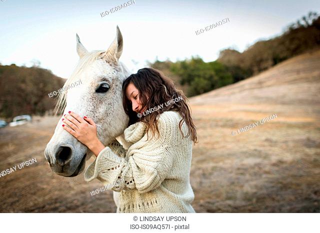Young woman leaning against and petting white horse in field