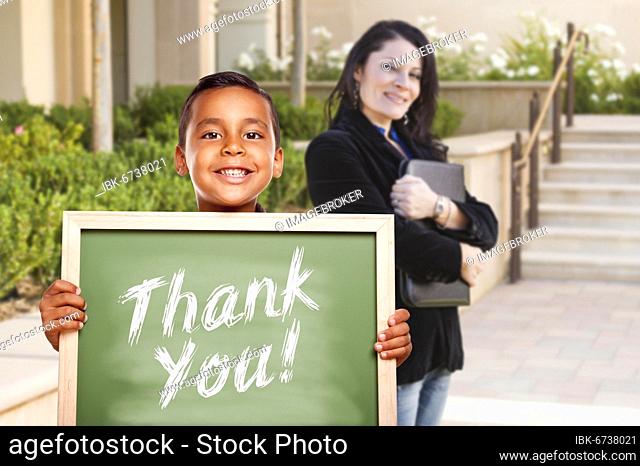 Happy hispanic boy holding thank you chalk board outside on school campus as teacher looks on