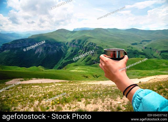 First-person view of a girl's hand holding a plastic cup of tea against a plateau of green hills and a cloudy sky in summer