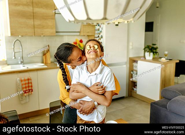Mother embracing son covering eyes with cucumbers in kitchen