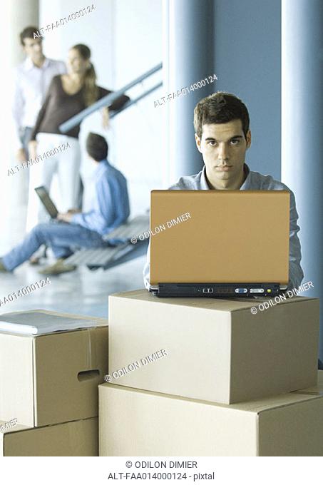 Man using laptop on stack of cardboard boxes, colleagues talking in background