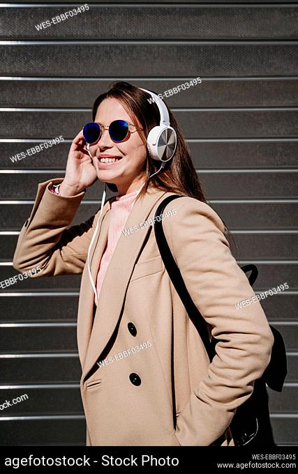 Young woman with headphones and sunglasses standing by wall