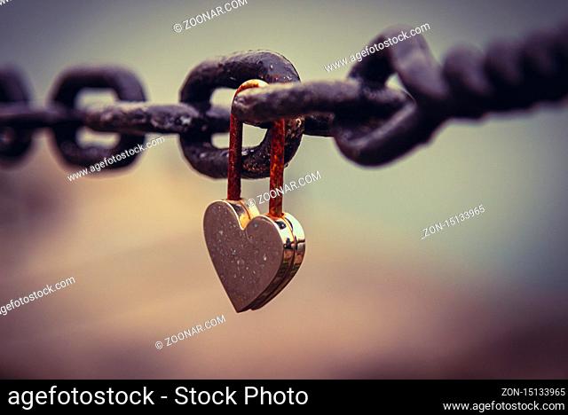 A romantic closed and rusty heart shaped padlock hanging from a chain beside a river with blurred cityscape in the background