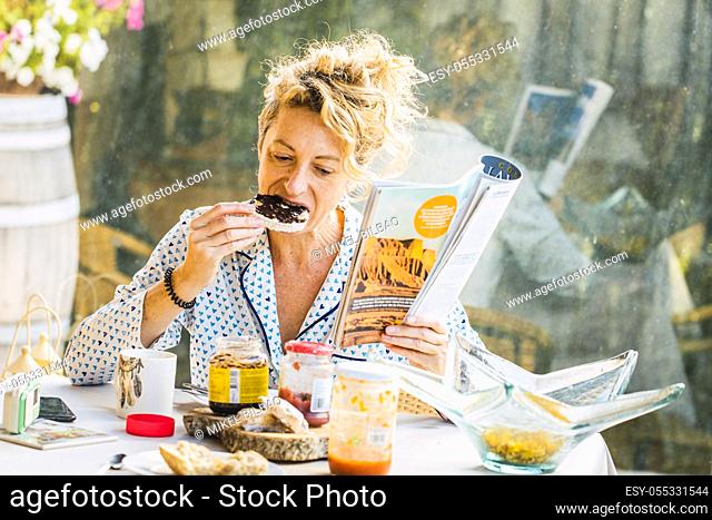 Blonde young mature woman in pyjamas at home in breakfast time, reading a magazine and eating a toast