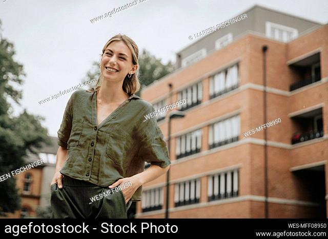 Happy young woman with hands in pockets in front of building
