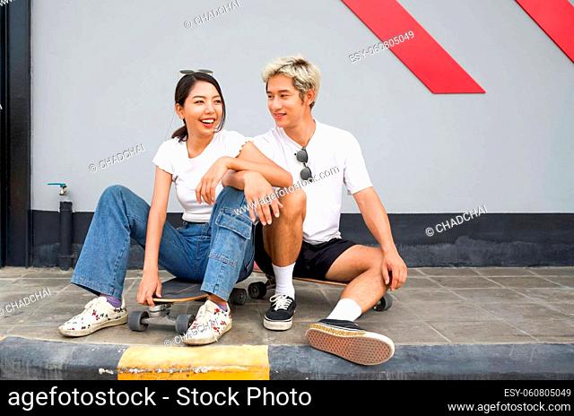 Asian couple resting after skateboarding. Asian woman in white T-shirt rest her arm at her knee. Young boyfriend sitting next to her
