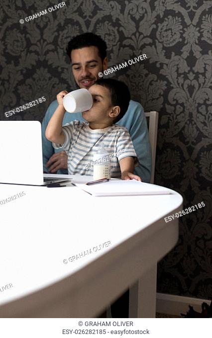 Young father is sitting at the table with his son on his lap. His son is copying what he has seen his dad doing and has his fathers mug to his mouth