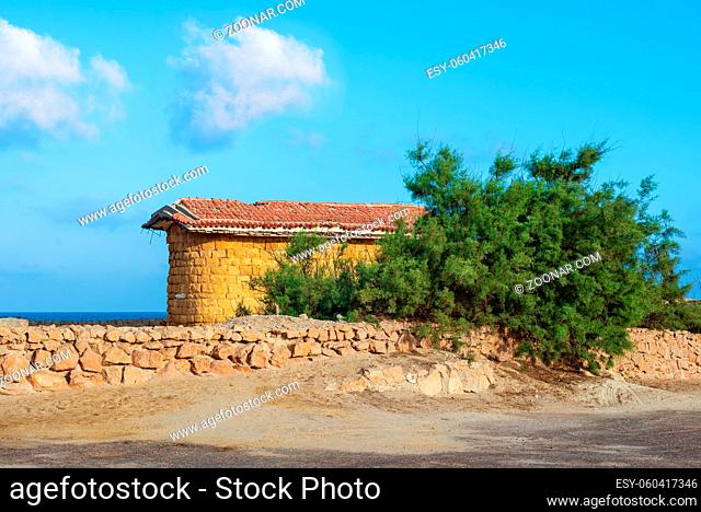 Abandoned building built of stone bricks and red tiled roof, short green tree and stone fence on background of partly cloudy sky and calm sea, Montaza Park