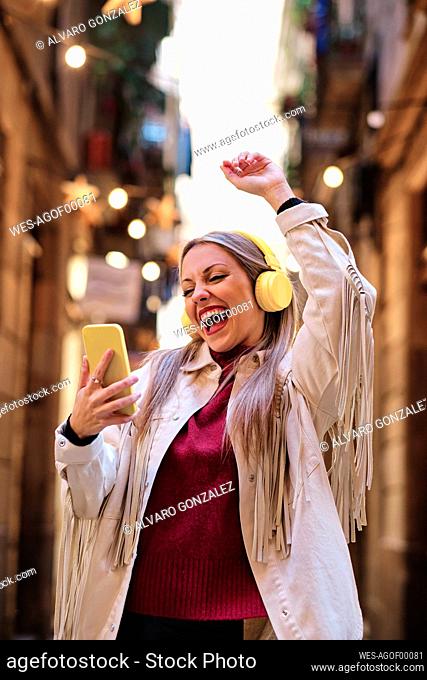 Cheerful woman with mobile phone dancing while listening music through headphones at alley