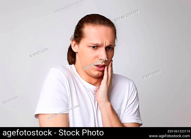 young unhappy man feeling pain, holding his cheek with hand, suffering from bad tooth ache on gray background