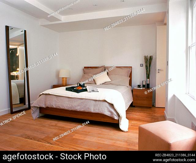 Details of a modern bedroom with a large full length mirror next to a double bed with two wooden side tables and a square seat in the foreground