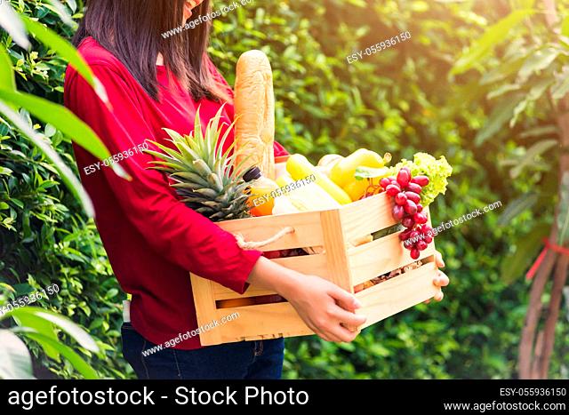 Portrait of Asian beautiful young woman farmer standing she smile and holding full fresh food raw vegetables fruit in a wood box in her hands on green leaves...