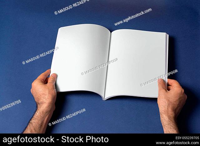 Male hands holding an opened book-catalog with blank pages on blue background, mock-up series template ready for your design, pages selection path included