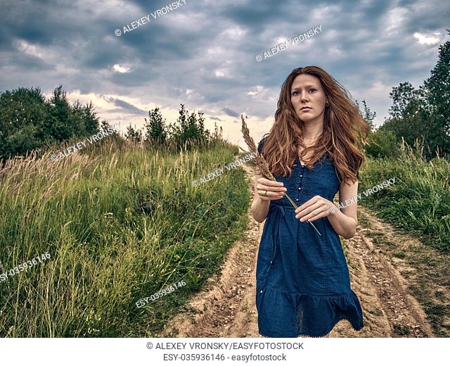 A girl with red long hair strolls along the sandy road along the forest lake. The girl's hair develops in the wind. The girl is dressed summer dress above the...