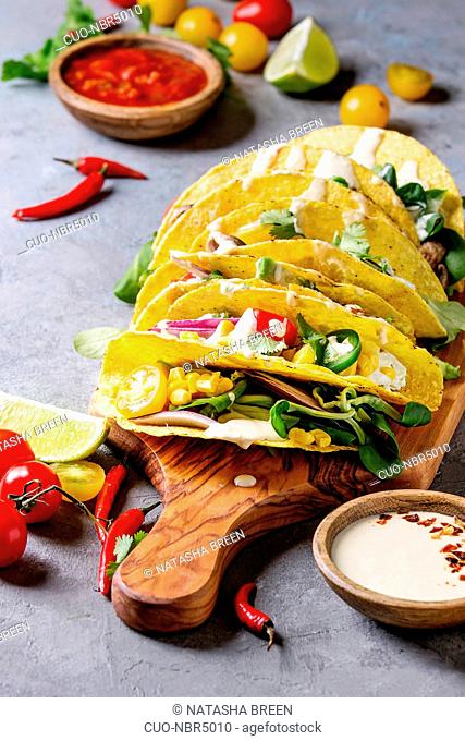 Variety of vegetarian corn tacos with vegetables, green salad, chili pepper served on wooden cutting board with tomato and cream sauces with ingredients above...