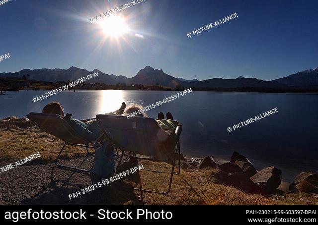dpatop - 15 February 2023, Bavaria, Füssen: Two women enjoy the sunshine on deck chairs on the shore of the Hopfensee lake on the edge of the Alps