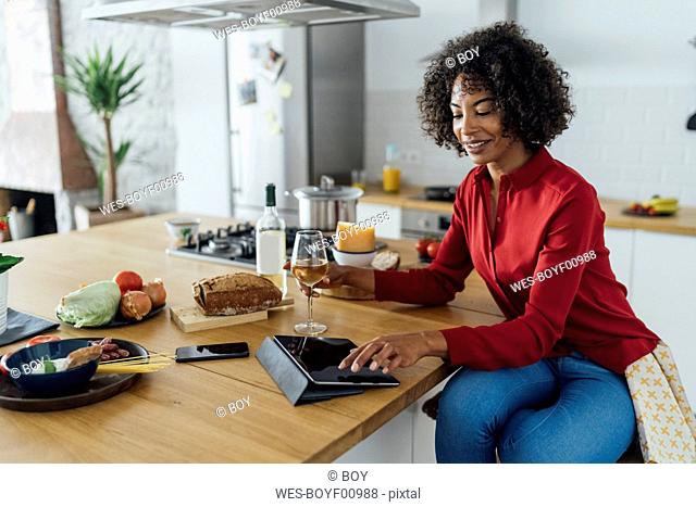 Woman sitting in kitchen with a glass of white wine, using digital tablet