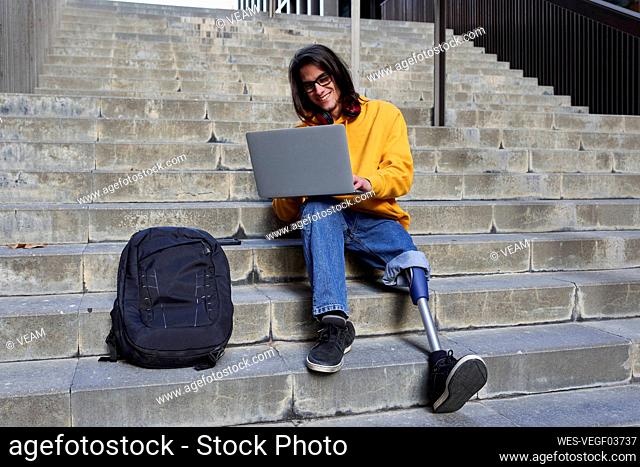 Smiling man with leg prosthesis using laptop while sitting on steps