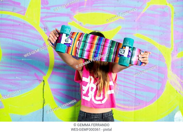 Teenage girl standing in front of wall with graffiti hiding behind colorful skateboard