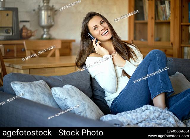 beautiful young smiling woman in white sweater talking on the phone on a gray sofa