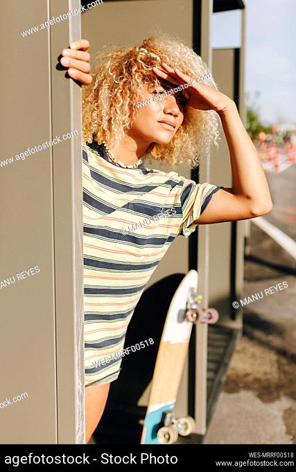 Blond Afro woman shielding eyes while looking away by metallic wall on sunny day