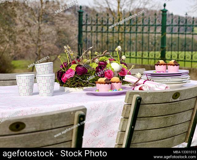 Easter breakfast table outdoors, in preparation, with coffee mugs, a stack of plates, muffins and a lush bouquet of flowers with ranunculus, feathers