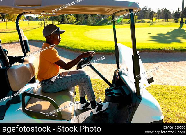 Side view of african american young man wearing cap riding golf cart at golf course in summer
